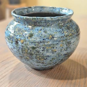 May include: A handmade ceramic vase with a wide, rounded body and a slightly flared rim. The vase features a speckled glaze in shades of blue, green, and gold. The interior is dark. This decorative piece is suitable for home decor.