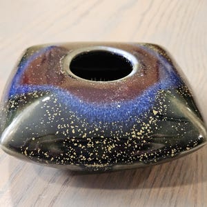 May include: A square-shaped, ceramic vase with a dark, glossy finish. The vase features a central opening and is adorned with a blend of blue, red, and gold speckles, creating a unique decorative piece.