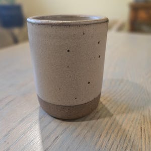 May include: A speckled ceramic tumbler with a light beige upper half and a darker brown base. The cylindrical cup has a slightly textured surface and a simple, minimalist design. The cup is suitable for hot or cold beverages.