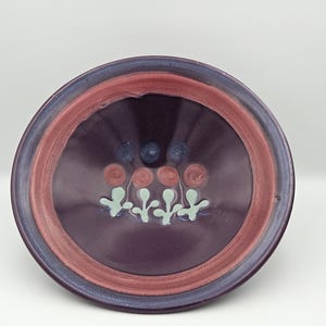 May include: A round ceramic bowl featuring a deep purple glaze with a wide terracotta-colored rim. The interior is decorated with a horizontal row of four pale green stylized floral motifs, topped by a row of four small terracotta-colored circles.