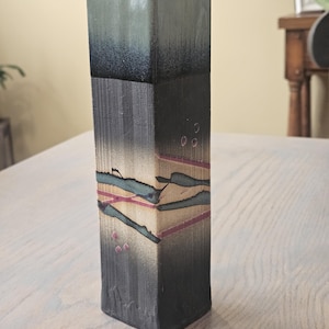 May include: A tall, rectangular ceramic vase with a dark, textured surface. The top is a gradient of dark blue to black. The body features horizontal bands of teal, pink, and beige. Small pink dots are scattered on the side. A decorative home accent.