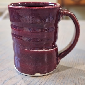 May include: A handmade ceramic mug in a deep burgundy color. The mug features a textured, ridged design around the body and a rounded handle. The interior and handle have a glossy finish, while the base is a lighter, unglazed color.