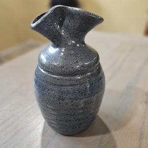 May include: A small, gray ceramic vase with a speckled glaze and a unique, ruffled opening.