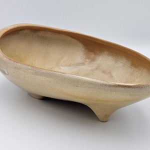May include: An oval-shaped ceramic planter with a smooth, glossy beige and tan glaze. The vessel features a deep, open basin and is supported by three short, sturdy feet integrated into the base, creating a raised, elevated appearance.