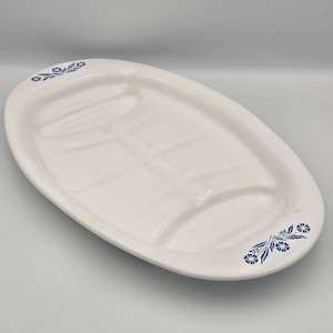 May include: An oval white ceramic serving platter featuring a recessed center with drainage grooves and a blue floral pattern on the two opposite ends. The platter has a smooth, glossy finish and a wide rim, designed for serving meat or poultry.
