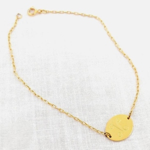 Könnte beinhalten: Ein goldfarbenes Kettenarmband mit einem feinen Gliederkettendesign und einem einzelnen ovalen Anhänger. Der Anhänger ist mit einem stilisierten, symmetrischen linearen Muster graviert. Das Armband verfügt über einen kleinen runden Verschluss.