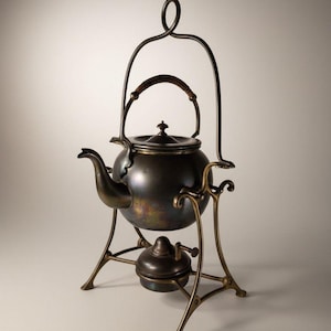 May include: Antique bronze tea kettle with a curved spout, a woven handle, and a decorative stand. The kettle has a dark patina and sits on a brass-colored frame with a burner below. The overall height is approximately 12 inches.