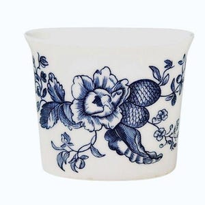 May include: A white ceramic cup featuring a blue floral pattern. The design displays a large central flower surrounded by detailed leaves, stems, and smaller blossoms. The cup has a slightly flared rim and a smooth, rounded body shape.
