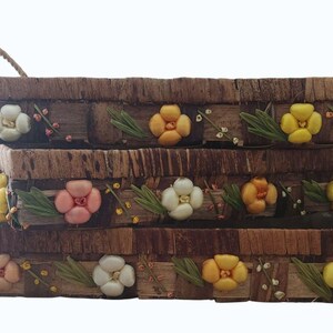 May include: A set of three rectangular brown woven trays with rope handles, featuring decorative three-dimensional flowers in shades of white, yellow, and pink, accented with green leaves and small sprigs arranged along the front exterior.
