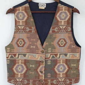 May include: A sleeveless vest featuring a multi-colored woven tapestry pattern in shades of rust, olive green, and muted blue. The garment has a V-neckline, four brass-toned buttons down the front, and a solid dark blue interior lining with a Clio label.