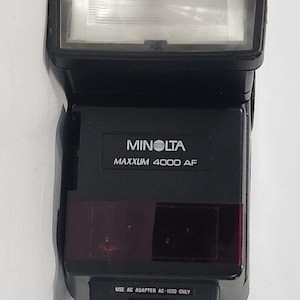 May include: A black Minolta Maxxum 4000 AF camera flash unit featuring a rectangular light head, a red sensor panel, and a base mount. The front displays the text Minolta Maxxum 4000 AF and the instruction Use AC Adapter AC-1000 Only.
