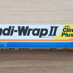Puede incluir: Caja rectangular de film plástico Handi-Wrap II. El embalaje es blanco con detalles azules, naranjas y amarillos. El texto "Handi-Wrap II" está en negro. Contiene 30,48 metros cuadrados de film.