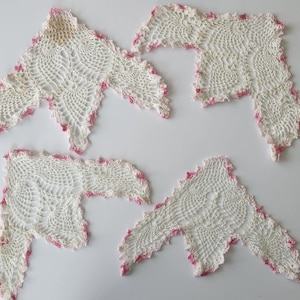 Può includere: Quattro sottobicchieri all'uncinetto bianchi fatti a mano con bordo smerlato e finiture rosa. I sottobicchieri sono disposti su una superficie bianca. L'uncinetto presenta un design traforato dettagliato.