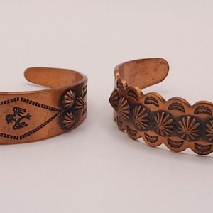 Pode incluir: Duas pulseiras de punho cor de cobre. Uma apresenta um padrão geométrico com formas de diamante e desenhos de raios de sol. A outra tem uma borda ondulada com padrões de estrelas repetidos. Ambas são abertas.