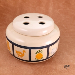 May include: Cream-colored ceramic flower frog with a lid featuring four holes. The base is decorated with a repeating pattern of yellow duck, pineapple, and rabbit illustrations. The flower frog is a decorative item.