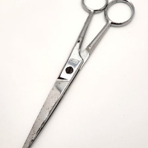 May include: A pair of silver-toned metal hair cutting shears featuring two finger rings, a curved finger rest, and a central screw mechanism. The blades are straight and pointed, showing signs of wear and minor surface imperfections on the metal.