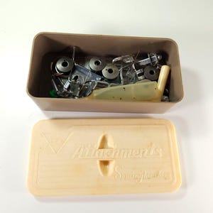 May include: A rectangular beige plastic storage box filled with various metal sewing machine attachments and bobbins. The matching lid is placed in front, featuring the embossed text Attachments Sewing Machine with a triangular logo design.
