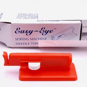 May include: An orange Easy-Eye sewing machine needle threader with a white button and a box with the text "Easy-Eye SEWING MACHINE NEEDLE THR". The box is white with blue text and a diagram of the threader.