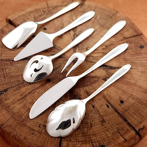 May include: A set of six silver-toned serving utensils including a wide shovel-shaped server, a triangular cake server, a slotted spoon, a two-pronged fork, a flat-bladed knife, and a decorative spoon with a scalloped, leaf-like bowl shape.