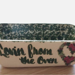 May include: A rectangular ceramic baking dish with a cream-colored exterior and interior. The exterior features the black text "Lovin from the Oven" and a painted heart motif with red flowers and green leaves. The interior has a green speckled pattern.