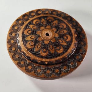 May include: A round wooden box lid featuring a dark brown pyrography design. The center displays a floral pattern with a small circular hole, surrounded by a border of repeating concentric circles etched into the light brown wood surface.