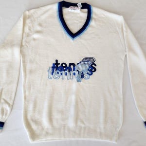 May include: A long-sleeve V-neck sweater in off-white featuring dark blue and light blue striped trim at the cuffs and neckline. The chest displays an embroidered graphic with the word tennis in layered blue text and a small tennis racket icon.