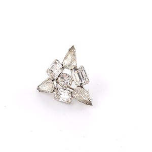 May include: A silver-toned brooch with a cluster of clear, faceted crystals in various shapes. The crystals include emerald cuts, pear shapes, and round stones, arranged in a triangular design. The brooch is set against a white background.