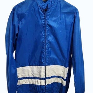 May include: A bright blue windbreaker jacket featuring a high collar, a full-length front zip, and two horizontal white stripes across the lower torso. The jacket has long sleeves with elastic cuffs and is displayed on a wooden hanger.