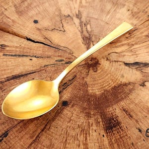 May include: A gold-colored spoon with a rounded bowl and a flat handle rests on a wooden surface. The spoon's polished surface reflects light, and the wood grain provides a contrasting texture. The spoon is likely used for serving or eating.