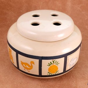 May include: A cream-colored ceramic flower frog with a rounded body and a removable lid featuring four circular holes. The base is decorated with a series of square panels outlined in dark blue, each containing illustrations of a duck, pineapple, and rabbit.