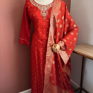 May include: A red salwar kameez suit with a matching dupatta. The suit has a V-neckline with detailed embroidery and small white polka dots. The dupatta has a gold patterned border and matching polka dots.