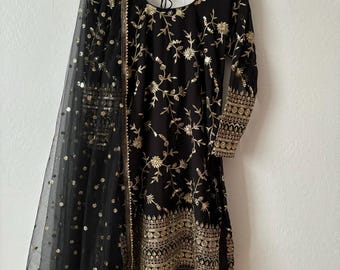 May include: Black embroidered tunic and matching trousers with a sheer black scarf. The tunic features gold floral embroidery and gold trim. The scarf is adorned with gold sequins. The outfit is hanging on a gold hanger.