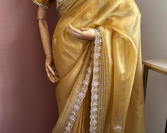 May include: A golden-yellow saree with intricate floral embroidery along the borders and neckline. The saree is draped on a mannequin, showcasing its flowing fabric and delicate details. The embroidery features pink and gold accents.