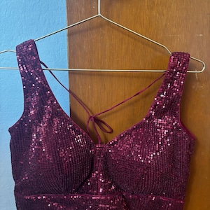 May include: A burgundy sequined top with a V-neck and crisscross straps. The sleeveless top is hanging on a gold hanger. The fabric is covered in small, reflective sequins, creating a shimmering effect. The top is a deep shade of red.
