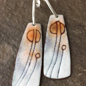 May include: A pair of dangle earrings with a white enamel background and a brown and orange abstract design. The earrings have silver hooks.