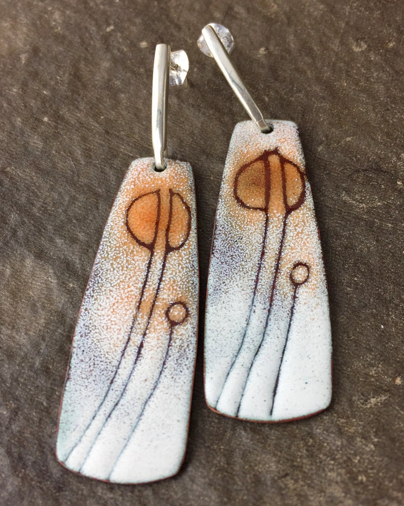 May include: A pair of dangle earrings with a white enamel background and a brown and orange abstract design. The earrings have silver hooks.