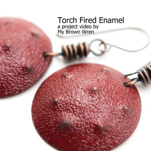 May include: A pair of red enamel earrings with a textured surface and copper wire accents. The earrings are shaped like small discs with a raised center and are attached to silver hooks.