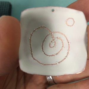May include: A white square pendant with a copper-colored spiral design and a small circle. The pendant has a hole at the top for hanging.