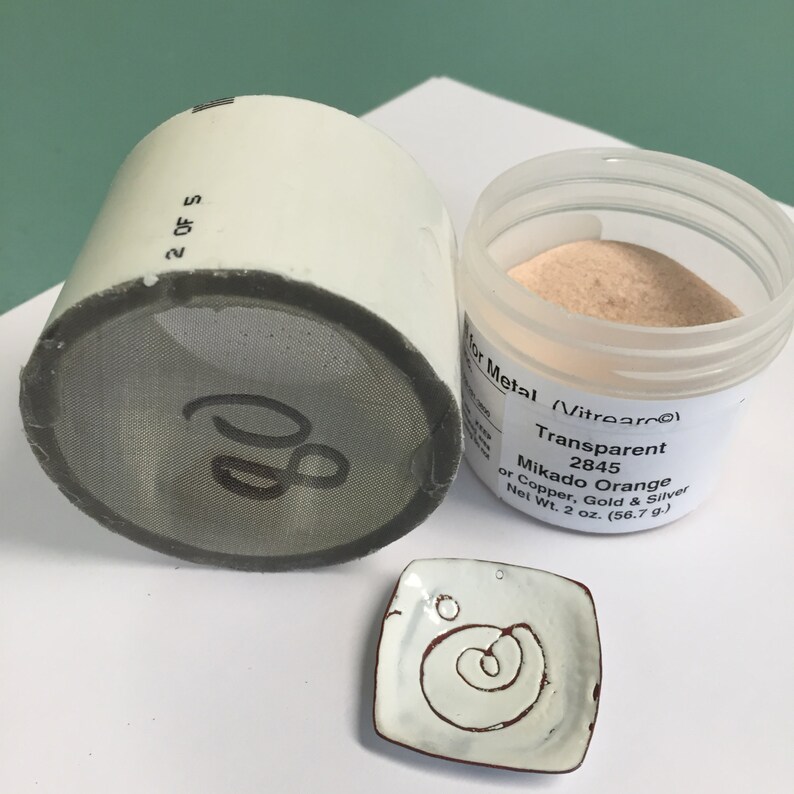 May include: A small white jar with a clear lid containing a light orange powder. The jar is labeled "Vitrearc Transparent 2845 Mikado Orange for Copper, Gold & Silver Net Wt. 2 oz. (56.7 g)".
