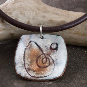 May include: A square pendant necklace with a white, brown, and orange abstract design. The pendant is made of ceramic and is attached to a brown leather cord.
