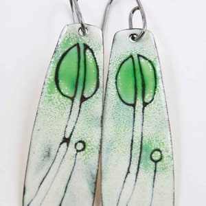 May include: A pair of dangle earrings with a white enamel background and green enamel accents. The earrings feature a stylized tree design with a thin black outline.