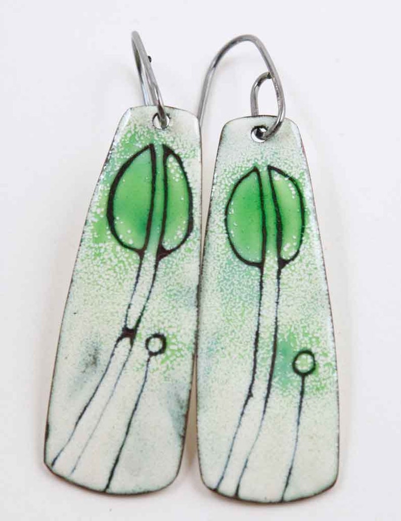May include: A pair of dangle earrings with a white enamel background and green enamel accents. The earrings feature a stylized tree design with a thin black outline.