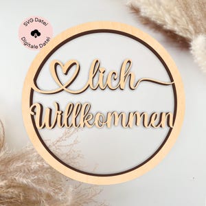 Pode incluir: Uma placa redonda de madeira com a palavra alemã "Willkommen" (Bem-vindo) em uma fonte decorativa. A placa tem um elemento de design em forma de coração e é cercada por uma borda marrom escura. A placa também inclui o texto "SVG Datei Digitale Datei" em um círculo rosa.