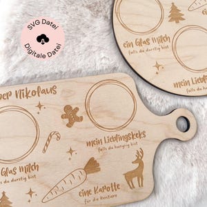 Op de afbeelding: Twee houten serveerplanken met gegraveerde vakantieontwerpen. E&eacute;n plank is rechthoekig met een handvat, met een peperkoekmannetje, zuurstok, wortel en rendier. De andere is rond met een kerstboom en een wortel. Tekst bevat "Der Nikolaus" en "mein Lieblingskeks".