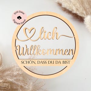Pode incluir: Uma placa redonda de madeira com as palavras "Willkommen" e "Schön, dass du da bist" recortadas. A placa tem um design de coração e um círculo menor com o texto "SVG Datei Digitale Datei".