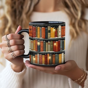Può includere: Tazza da caffè nera con un design a libreria. La tazza presenta file di libri colorati in tonalità di rosso, arancione, giallo e blu. La tazza ha un manico e un bordo neri, con piccole luci appese.