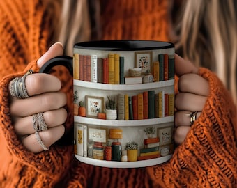 May include: A black-handled coffee mug featuring a bookshelf design with colorful books, framed art, and potted plants. The mug has a black rim and a white interior. The design includes various shades of red, yellow, green, and brown.