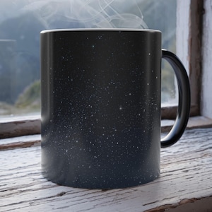 Könnte beinhalten: Eine schwarze Keramiktasse mit Sternennacht-Design. Die Tasse hat einen schwarzen Henkel und einen weißen Rand. Dampf steigt aus der Tasse auf, was auf ein heißes Getränk hindeutet. Die Tasse steht auf einer Holzfensterbank.