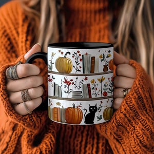 May include: White ceramic mug with a black interior and handle. The mug features a design of pumpkins, books, flowers, and a black cat. The mug is held by a person wearing an orange sweater and rings.