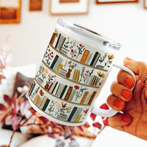 Op de afbeelding: Witte keramische mok met een ontwerp van boekenkasten vol boeken en bloemen. Het ontwerp bevat verschillende kleuren zoals groen, oranje en bruin. De mok heeft een wit handvat en een doorzichtig deksel.